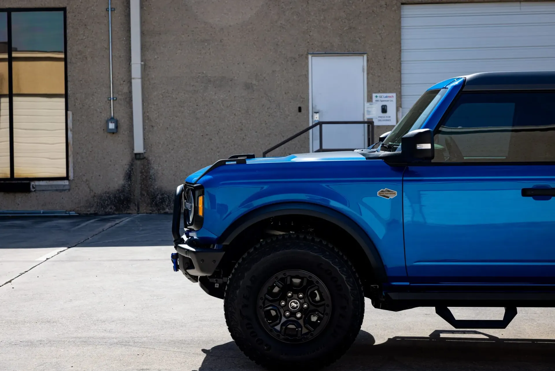 Blue Ford Bronco parked next to a building with a black roof and black wheels.