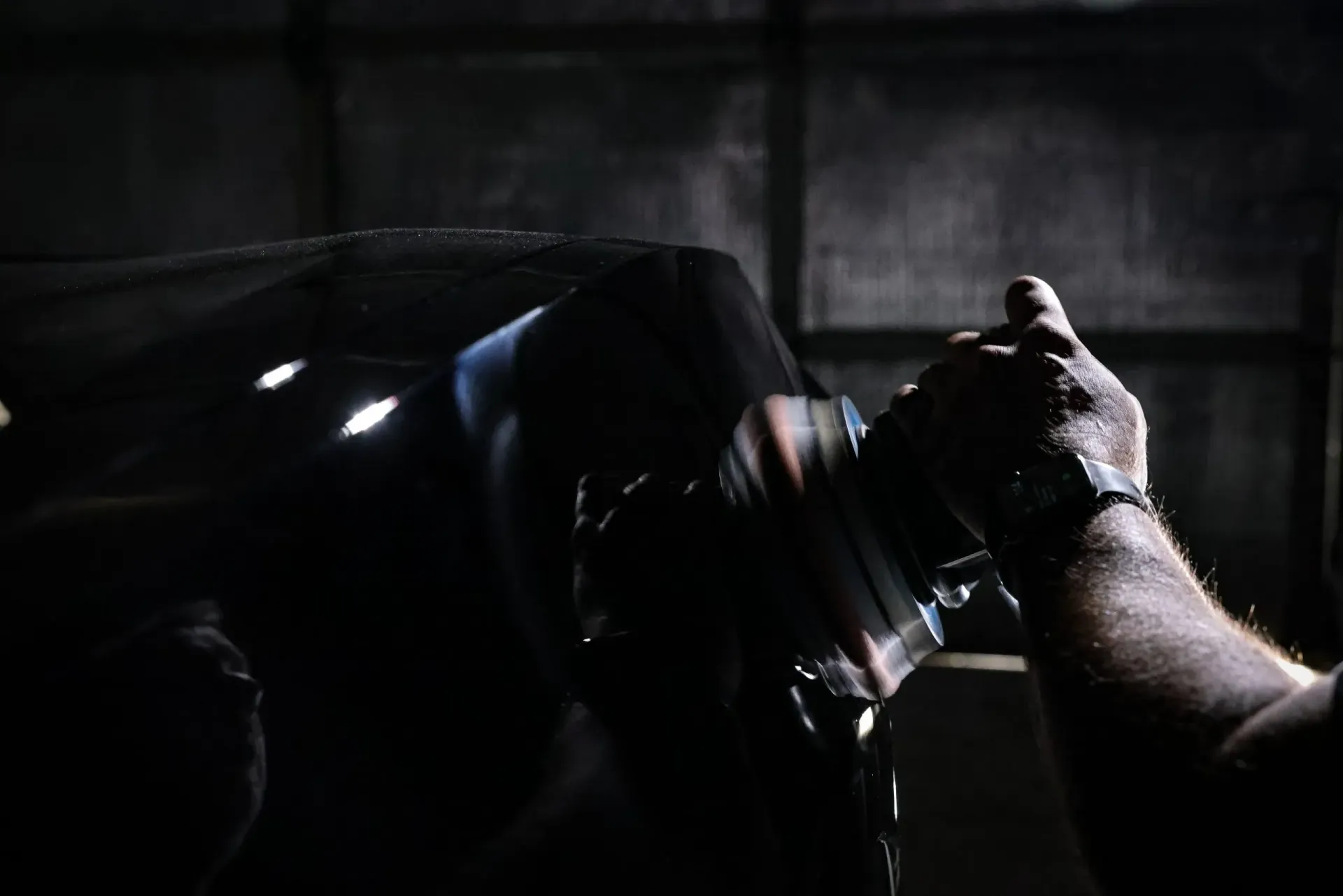 A person polishing a black car with a power tool in a dimly lit garage.