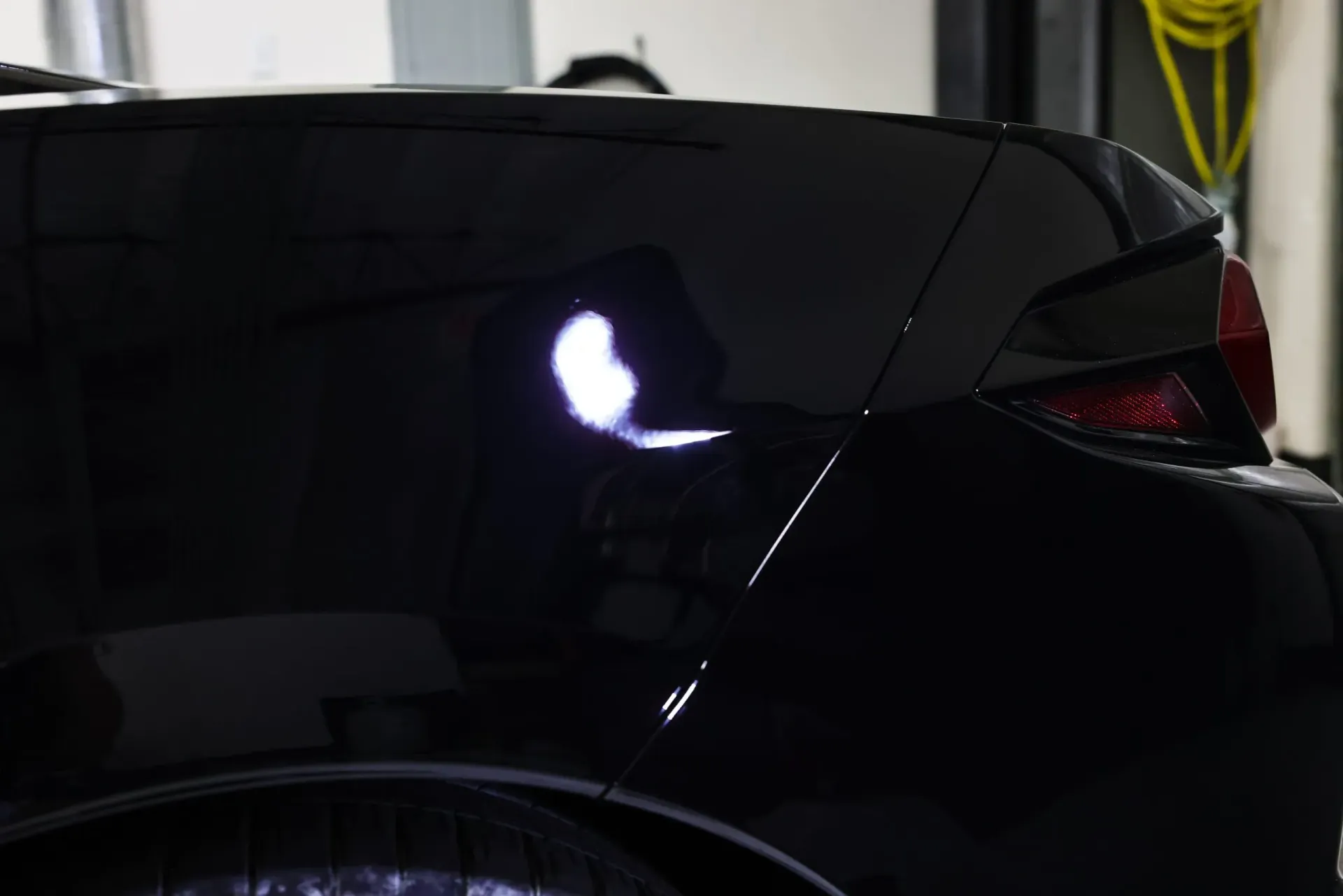Black car's rear quarter panel gleaming under bright lights in a shop; a close-up showing reflections.