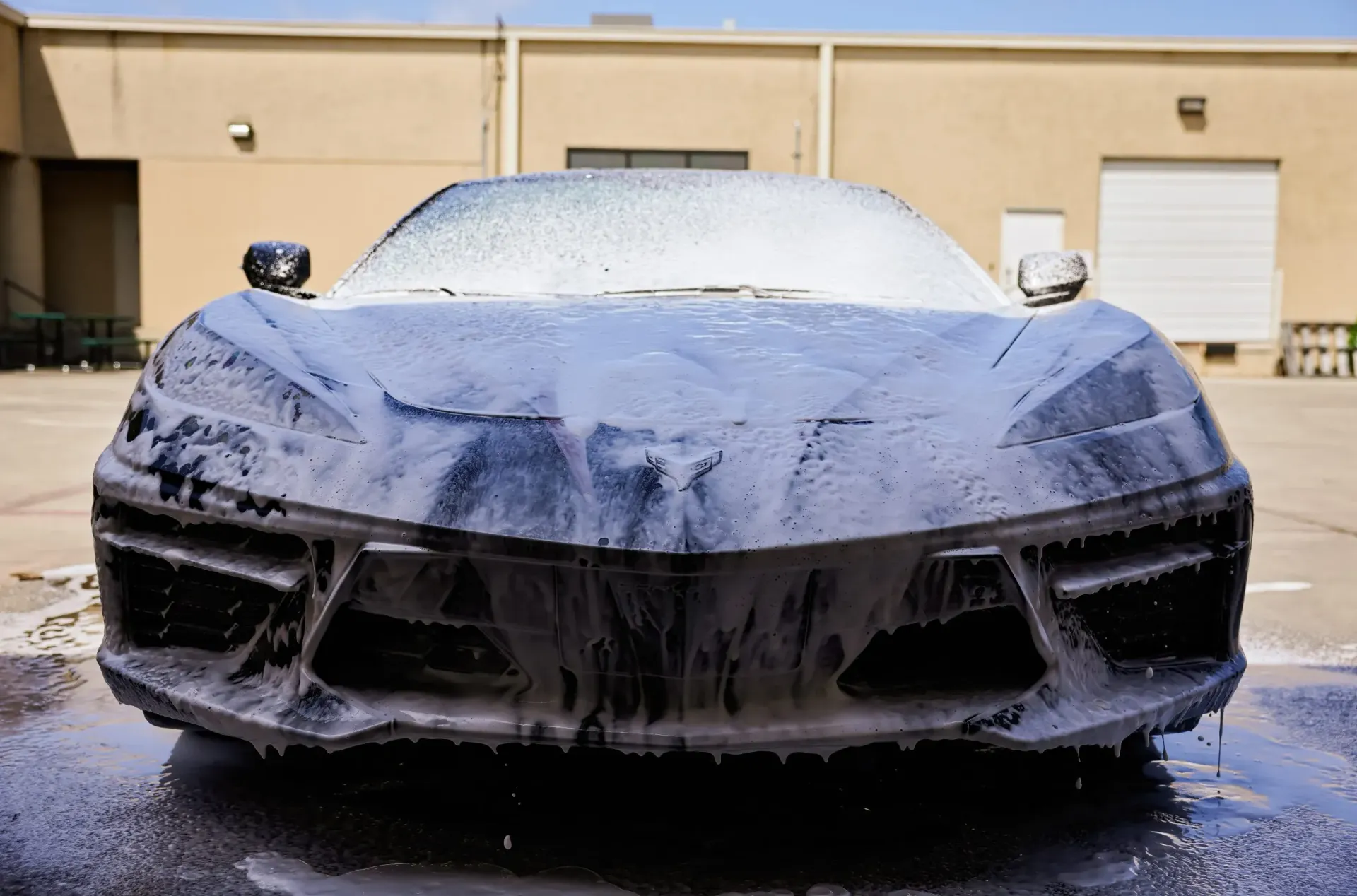 A black sports car covered in white foam during a car wash, outdoors.