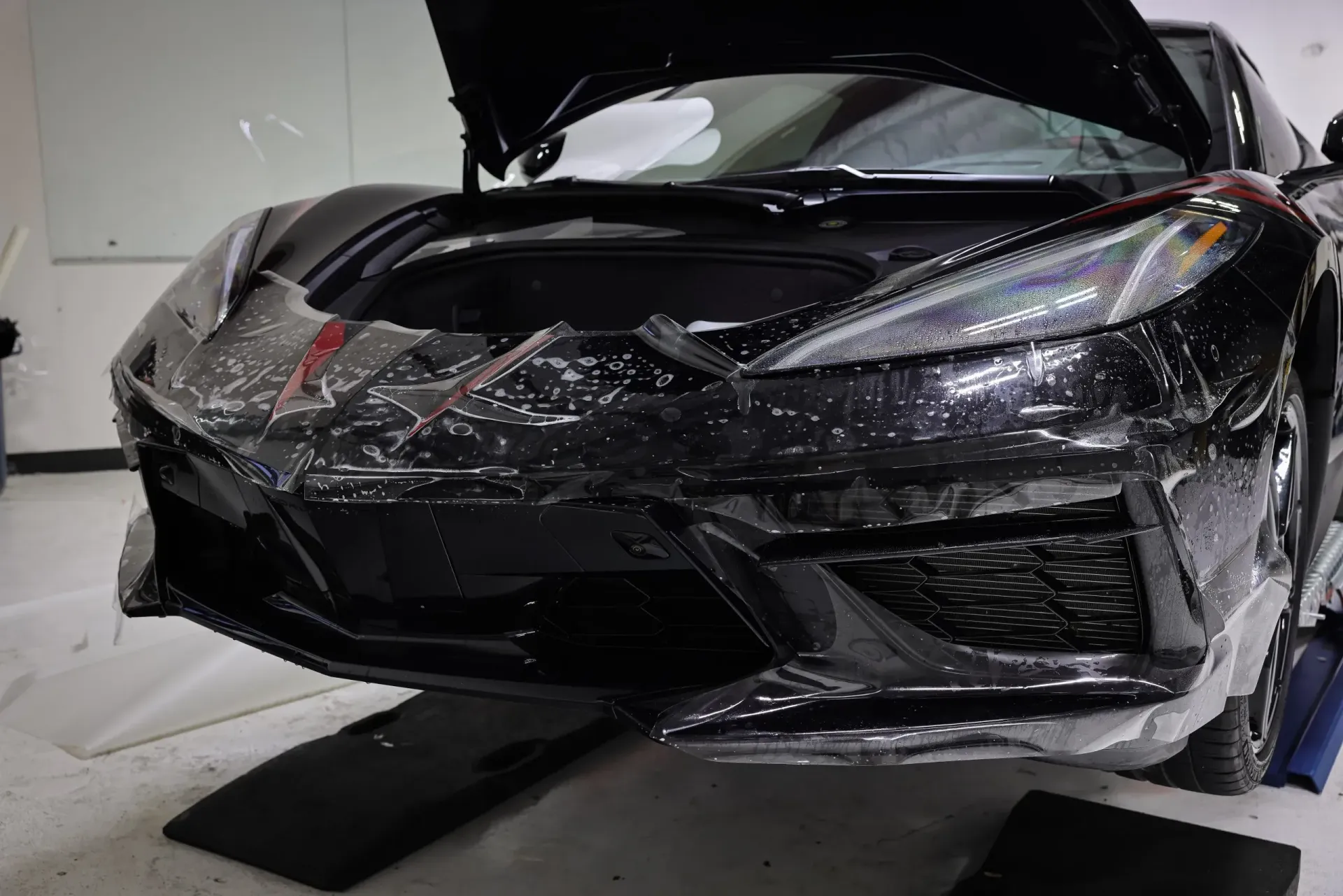 Black car with protective film applied to the front bumper. Garage setting, hood open.