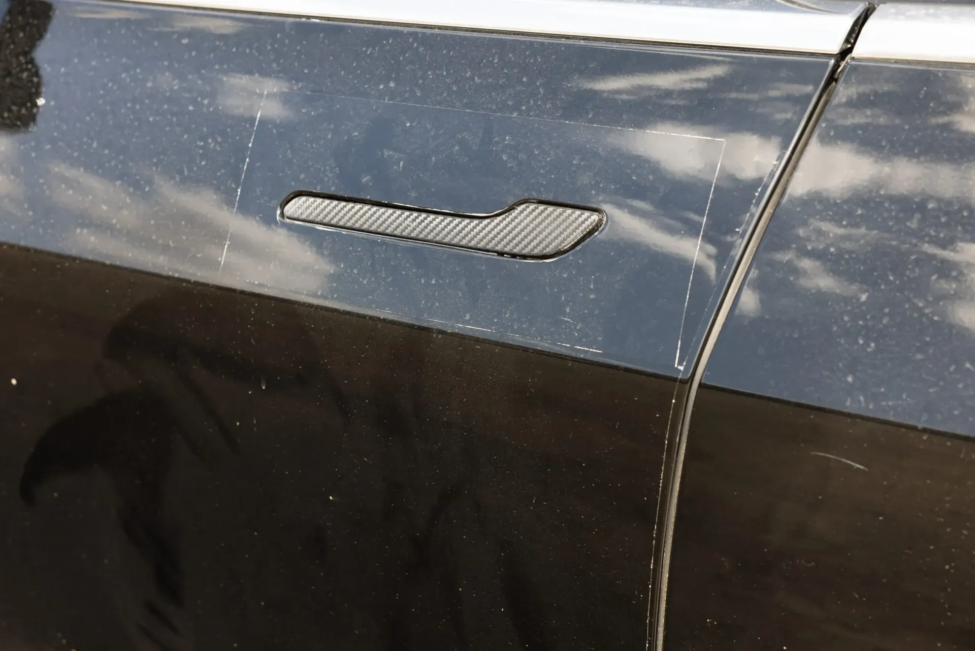 Close-up of a black car door with a carbon fiber door handle. The car has water droplets.