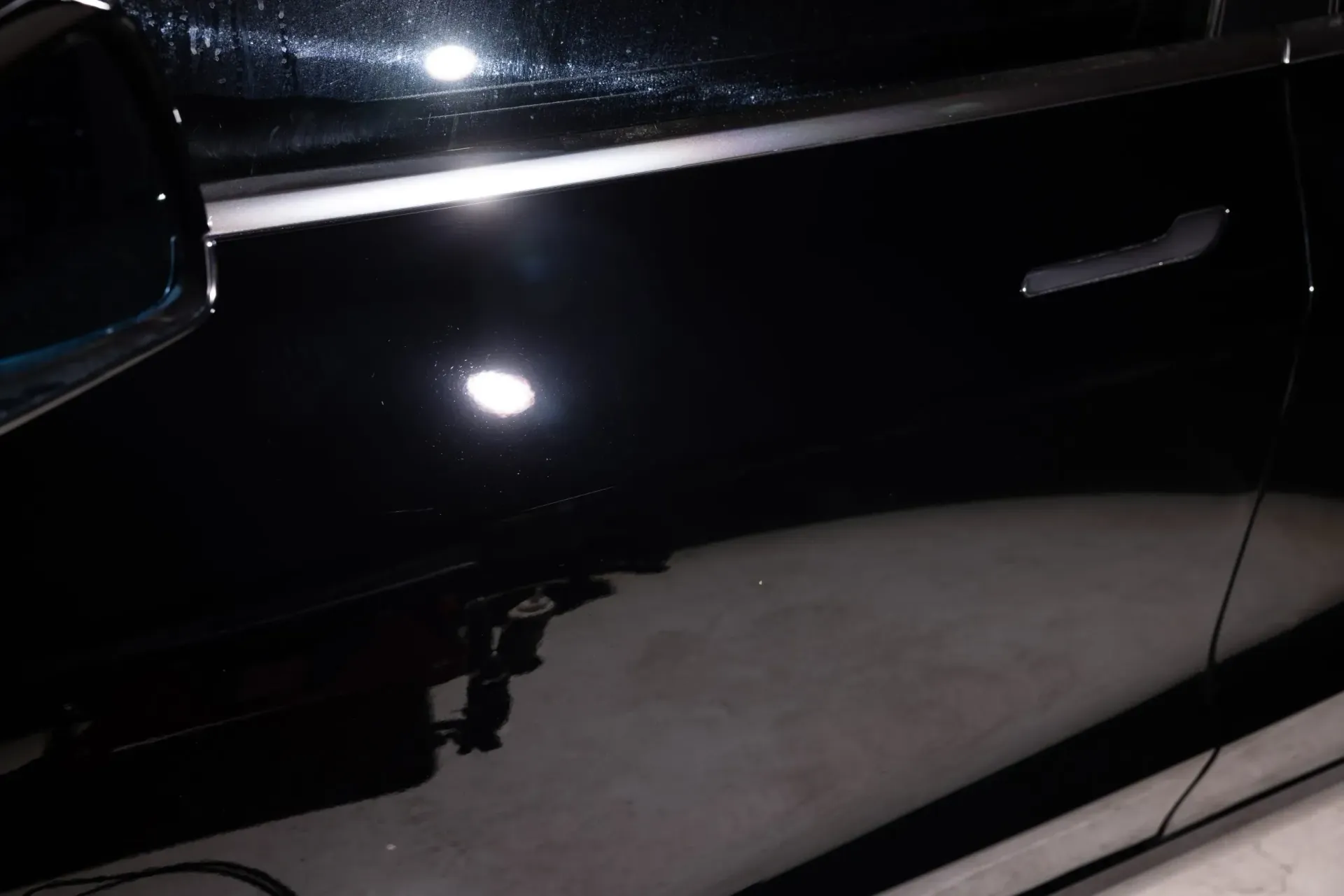 Black car door reflecting light, with handle visible, indoors.