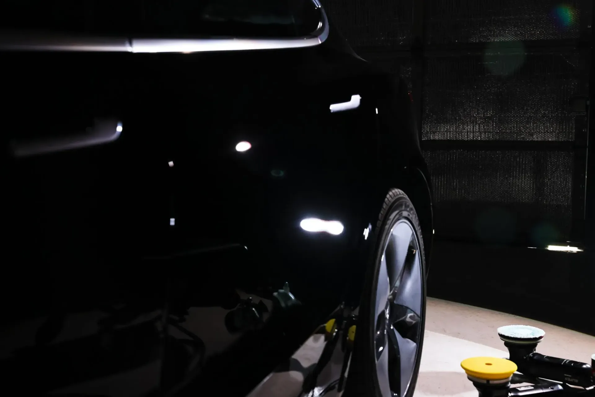 Black car with a shiny finish, the rear wheel is visible. Polishing equipment is in the foreground.