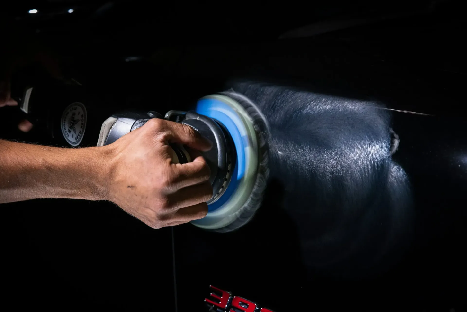 Person polishing a black car with a rotating buffer, highlighting the car's surface.