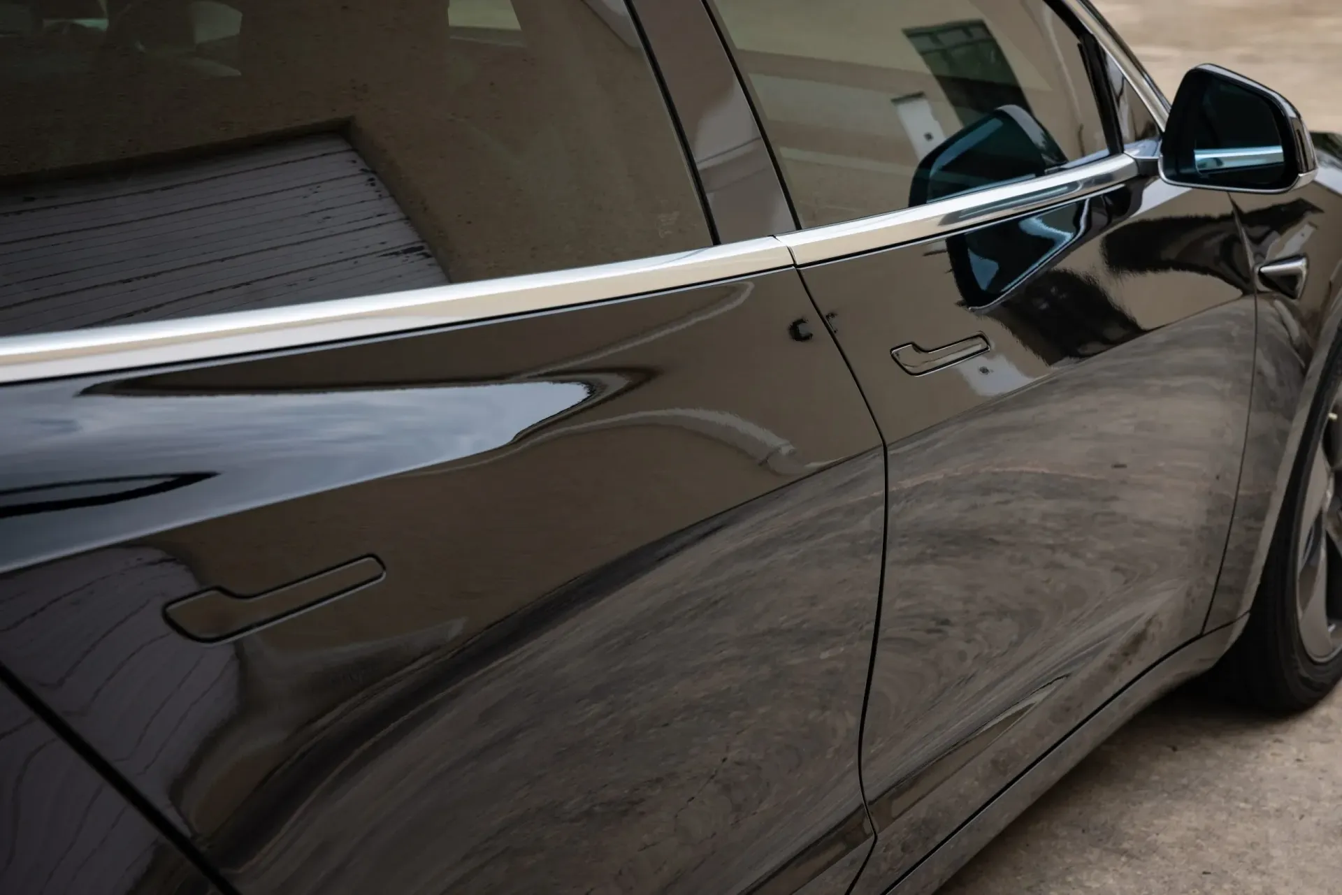 Black car with chrome accents, parked on a concrete surface. The Tesla logo is visible on the door.