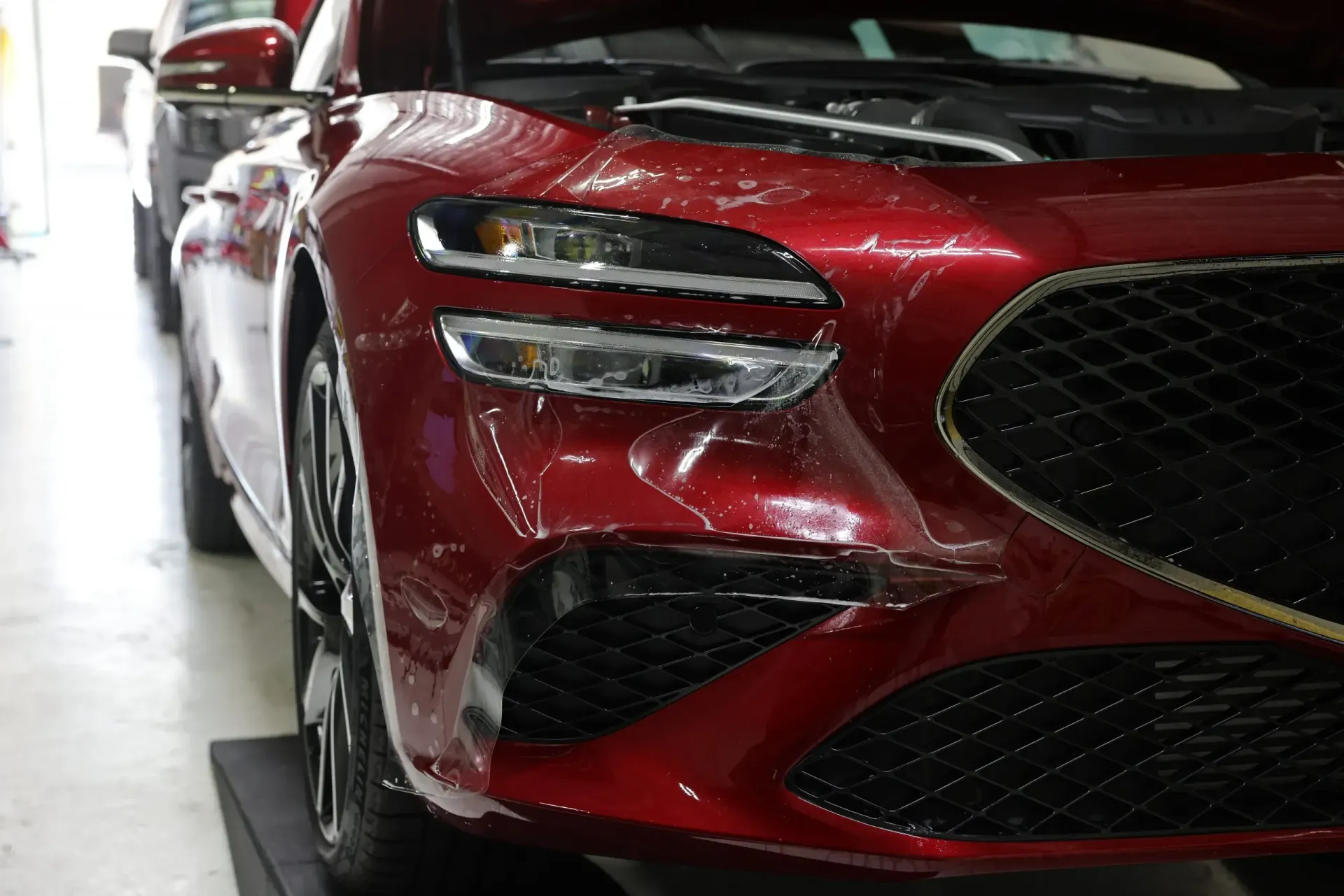 Red car with hood open, undergoing protective film application on its front.