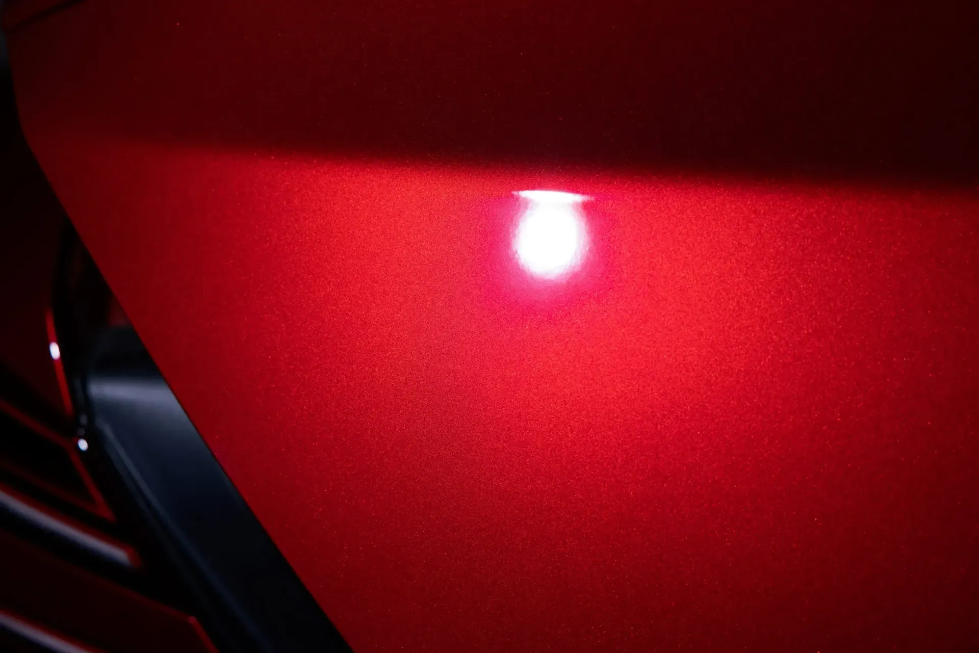 Close-up of a red car panel with a bright white reflection of a light, highlighting the paint's texture.