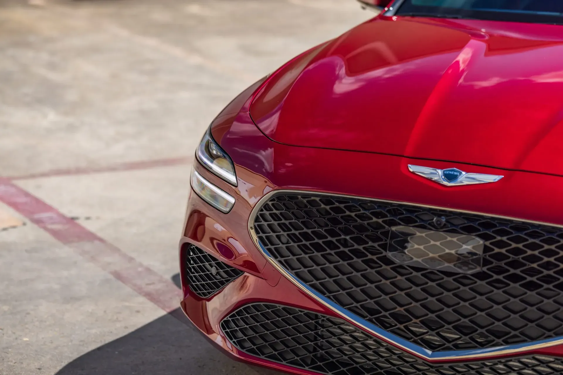 Red Genesis G70 car's front. Shiny hood, grill with Genesis logo. Outdoors, sunny.