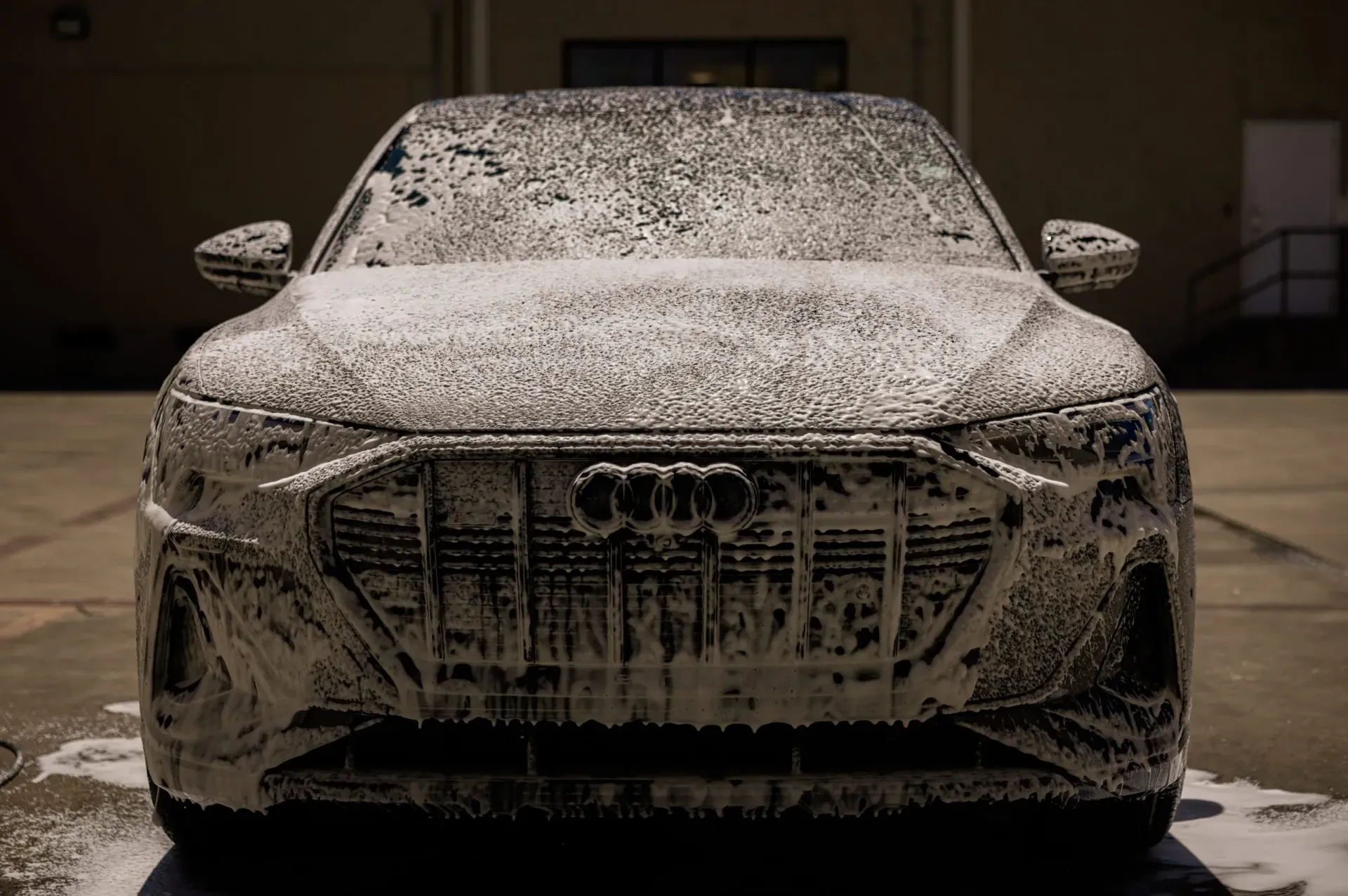 An Audi covered in white foam, viewed head-on in a sunny outdoor setting.