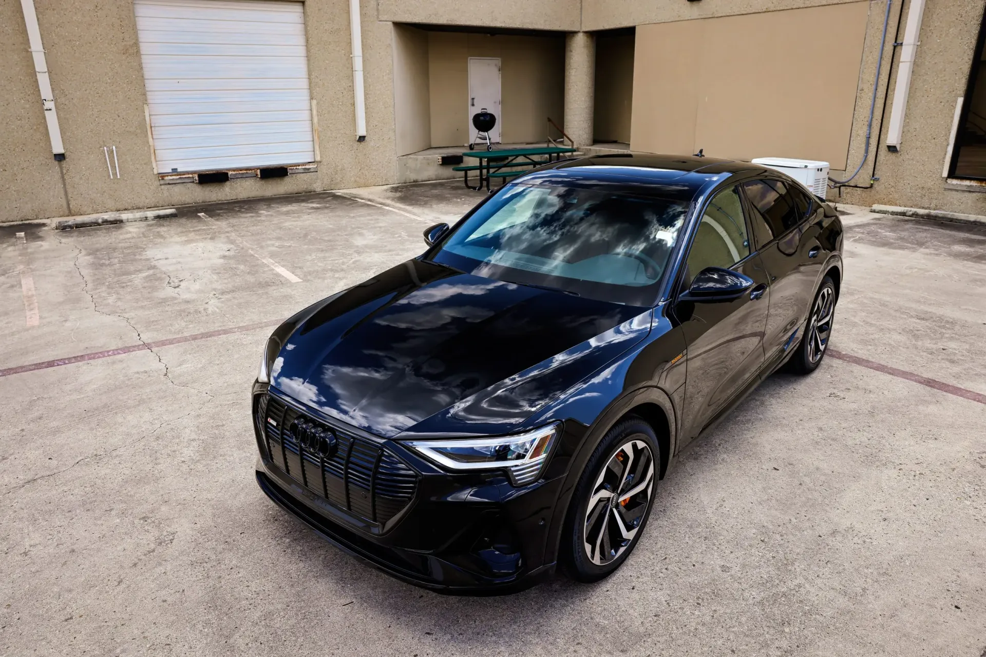 Black Audi e-tron electric car parked in an outdoor lot near a building, reflecting the sky.