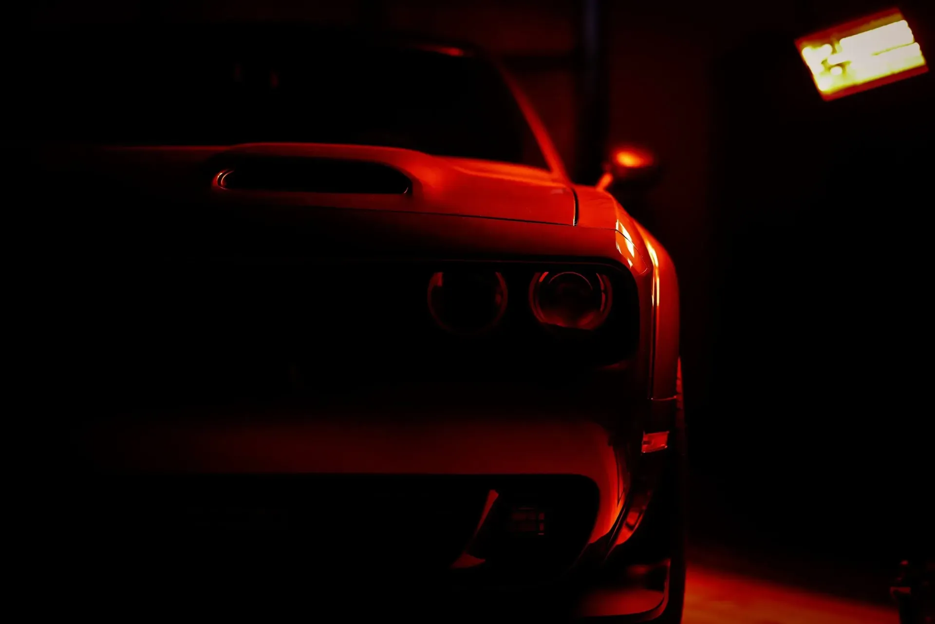 Red-lit front of a Dodge Challenger muscle car in a dimly lit garage.