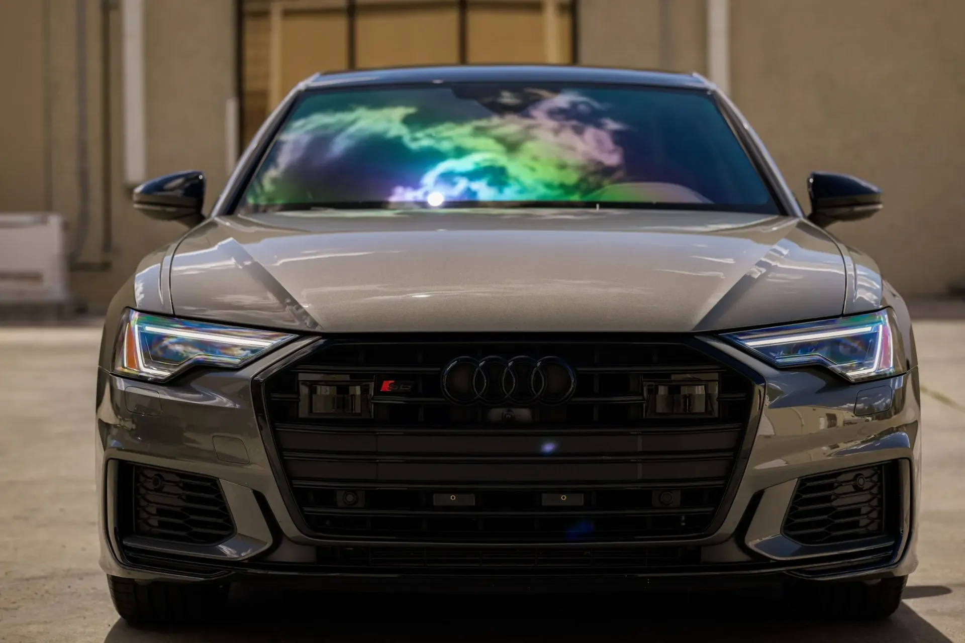 Gray Audi sedan with black accents; front view. Windshield reflects colorful light.