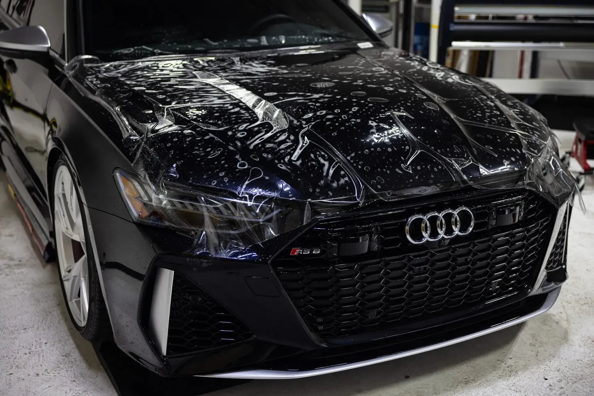 Black Audi RS6 with clear protective film on hood, in a garage.