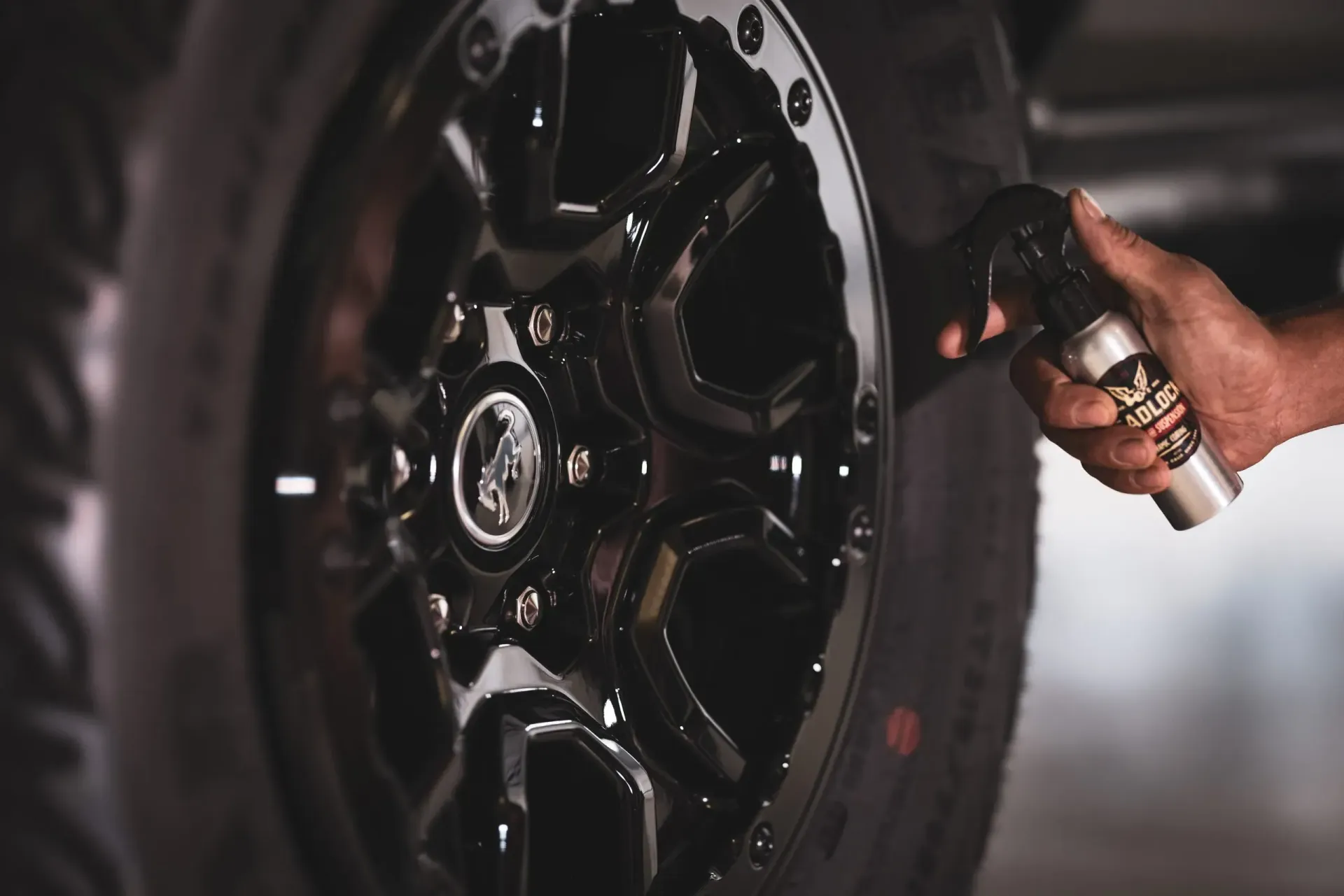 Person spraying a shiny black car wheel with a silver bottle, close-up shot.