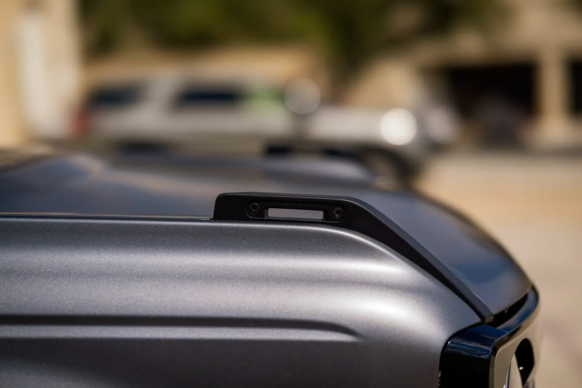 Close-up of a dark gray vehicle hood with a black accessory on top, blurred background with other vehicles.