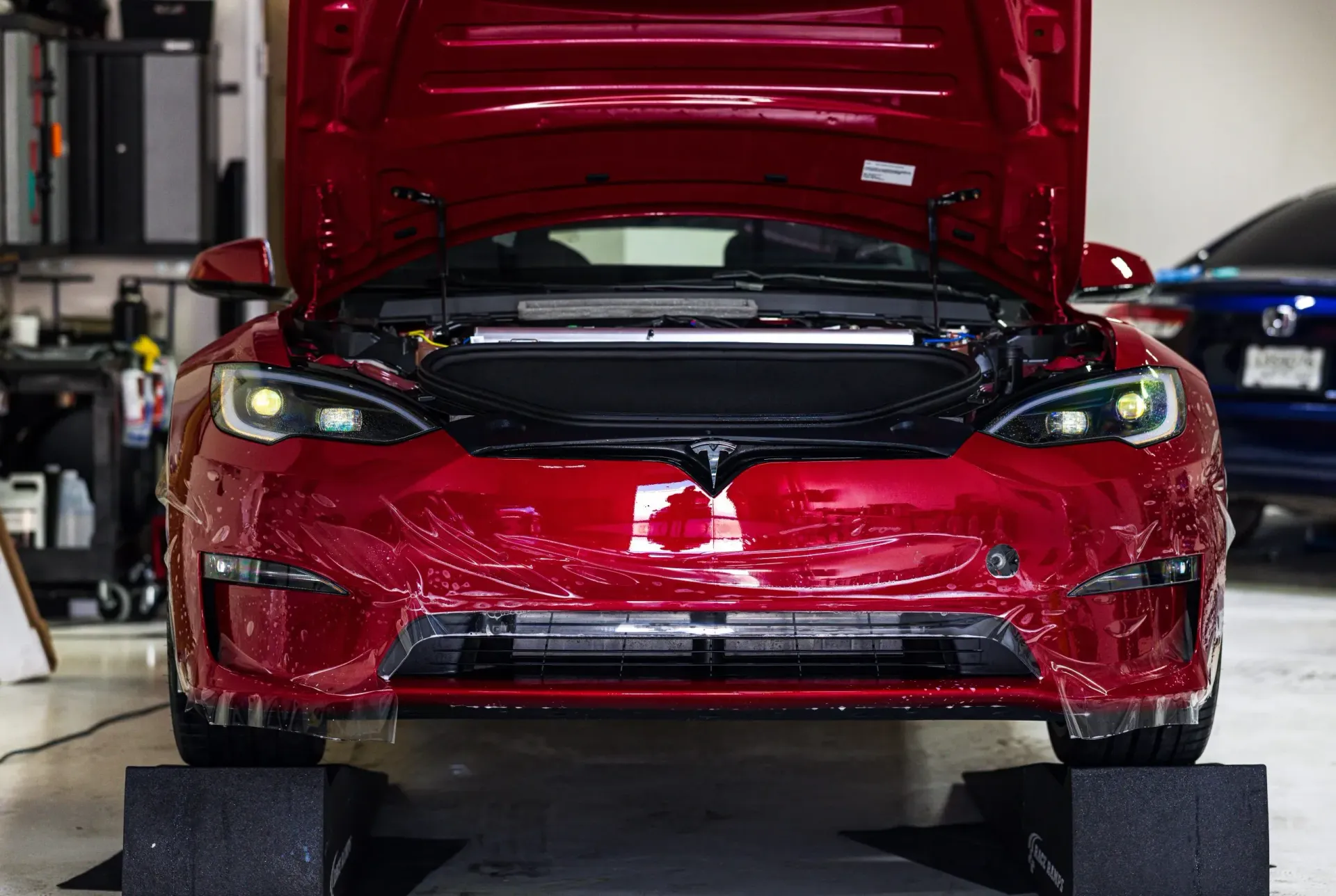 Red Tesla with open hood, undergoing repair or modification, in a garage.