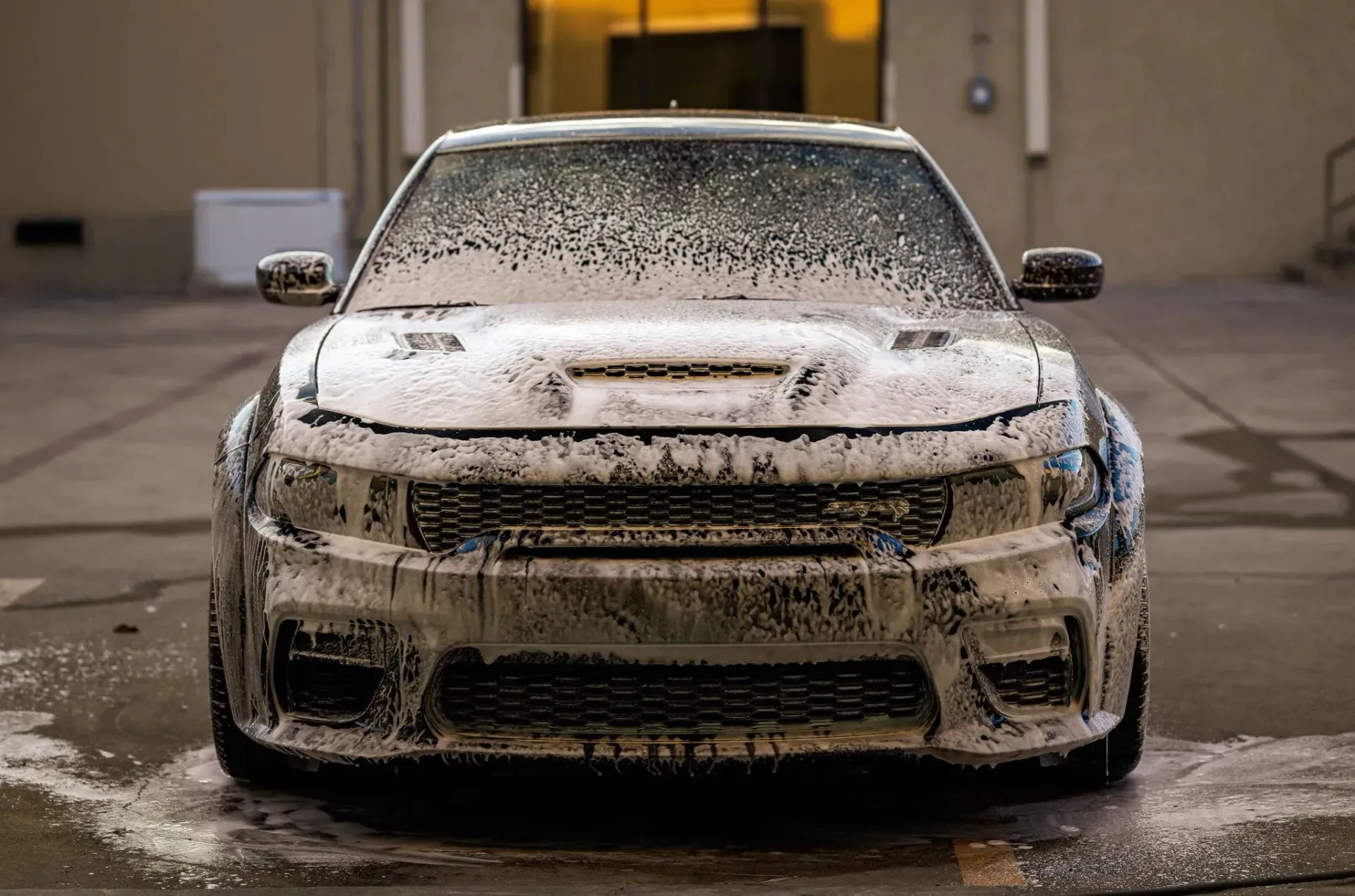Black car covered in white foam, being washed outside a building.