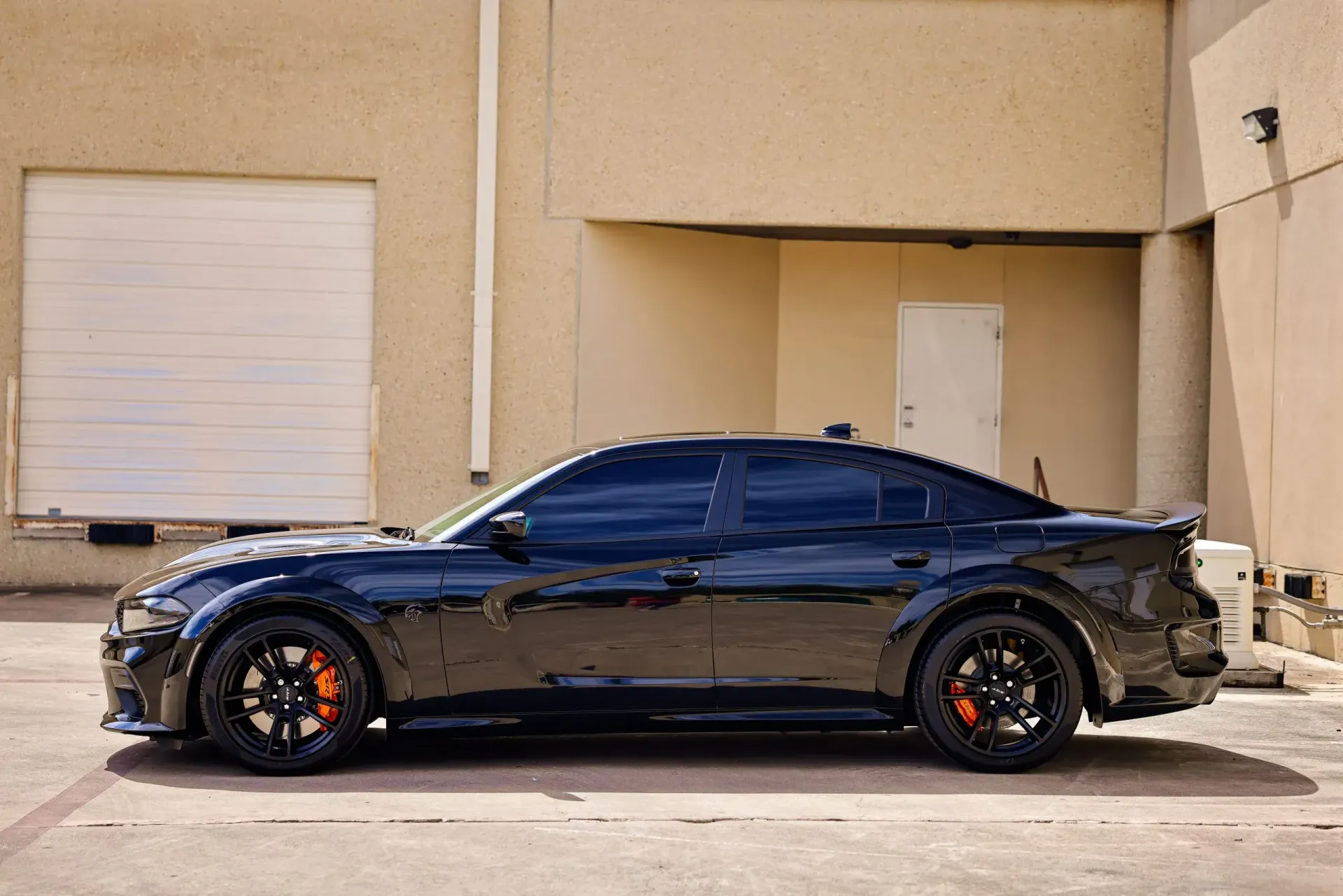 Black Dodge Charger with orange brake calipers parked in front of a tan building with a garage door.