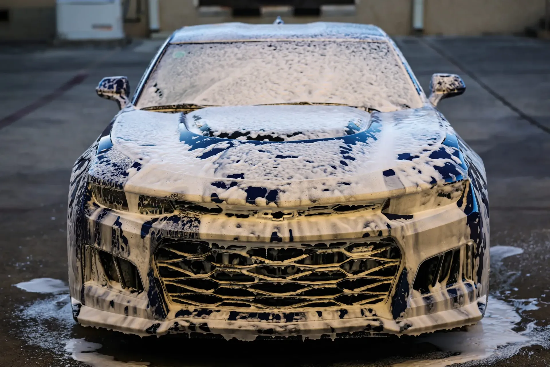 Blue car covered in white foam, presumably being washed outside.