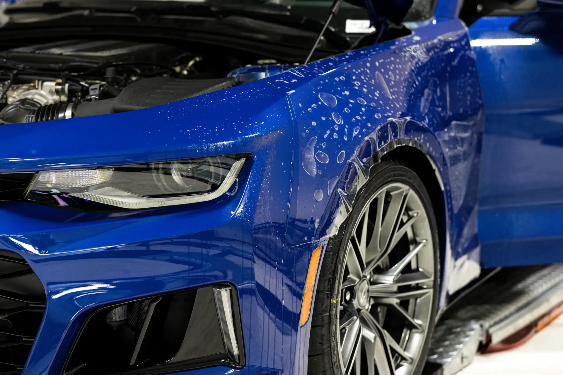 Blue sports car, close-up, wet. Hood open, detailing the front fender, headlight and tire.