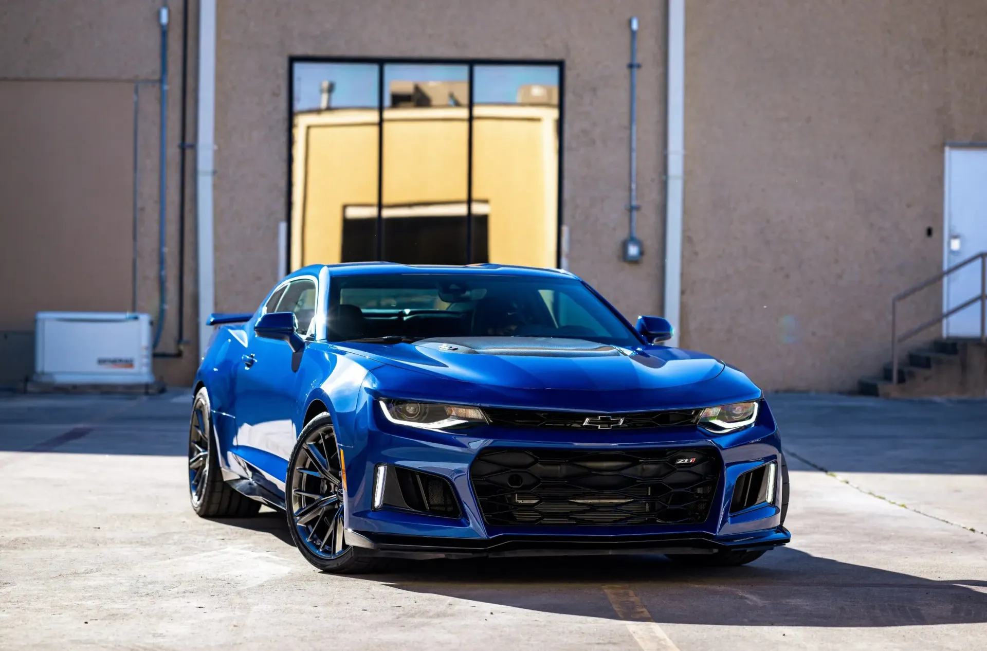 Blue Chevrolet Camaro sports car parked in front of a beige building with a large window.
