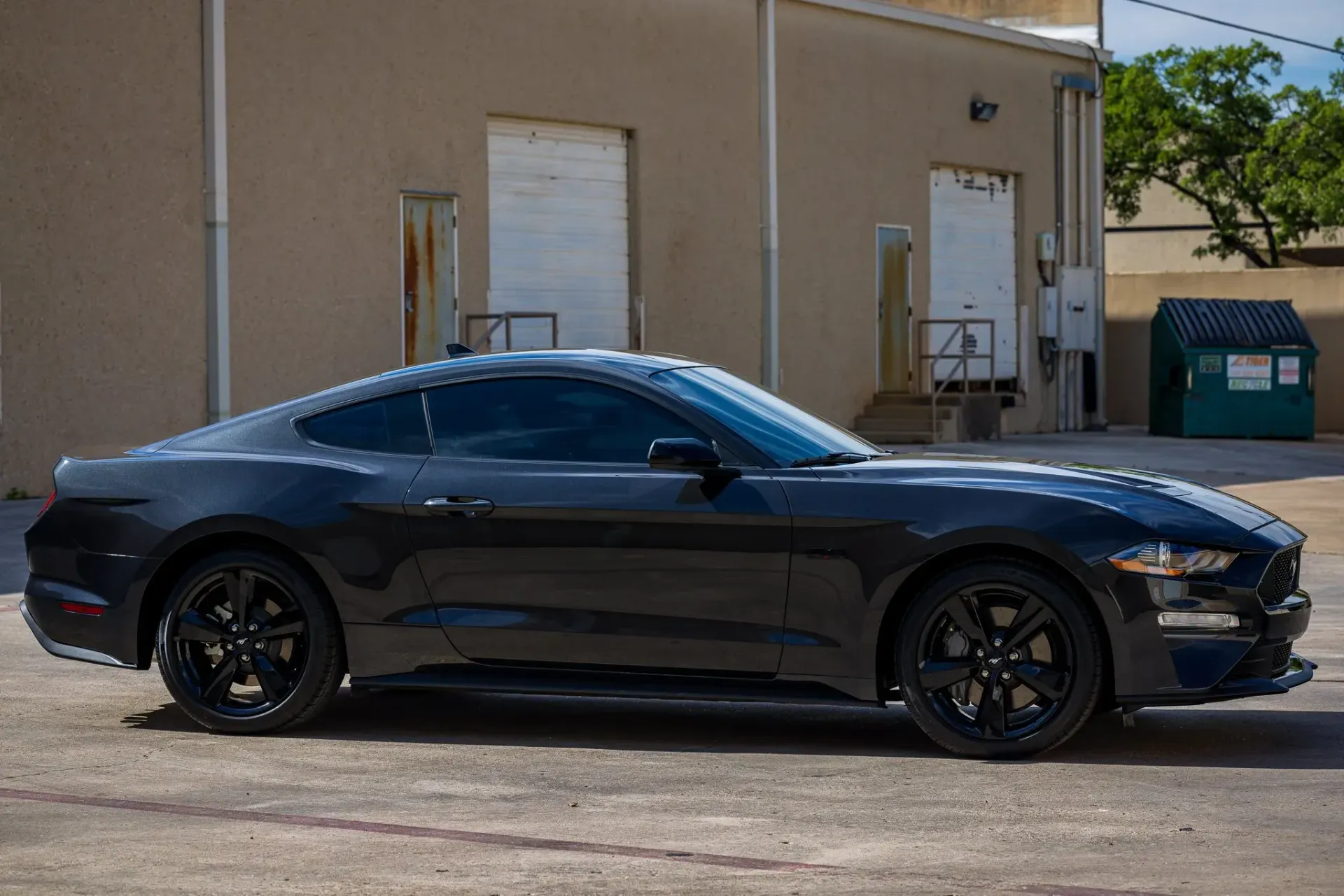 Black Ford Mustang parked in front of a tan building with white doors and a green dumpster.