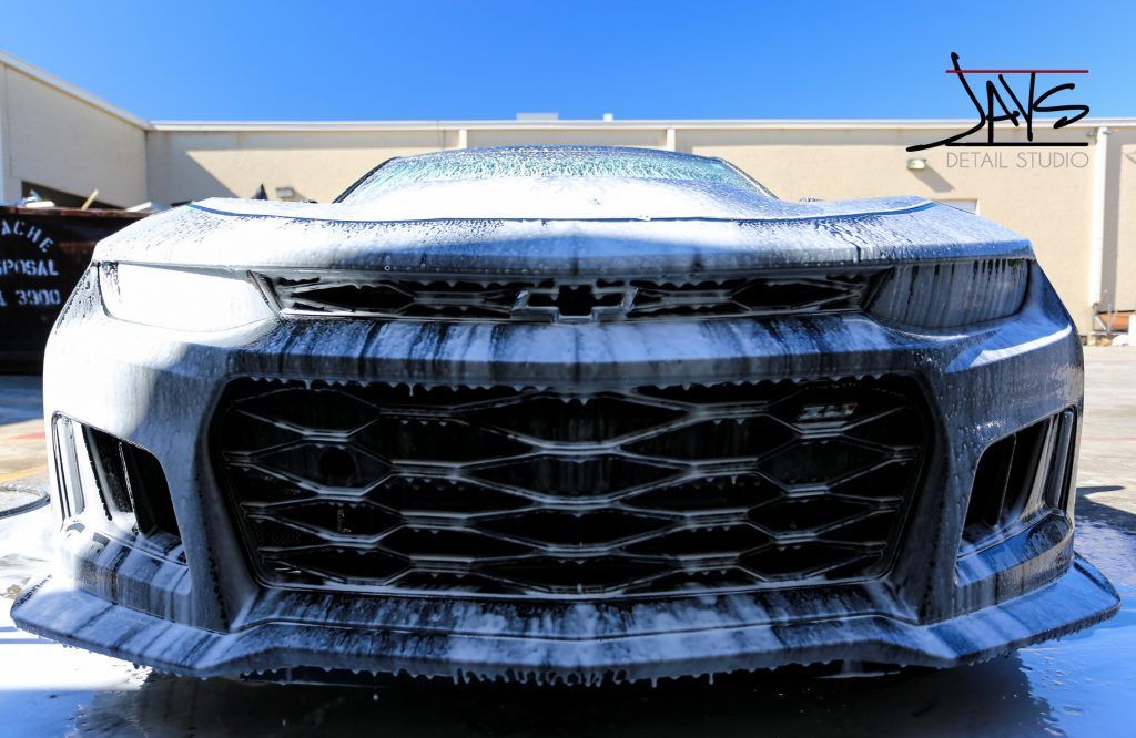 A black sports car covered in white foam, being washed outside a building.