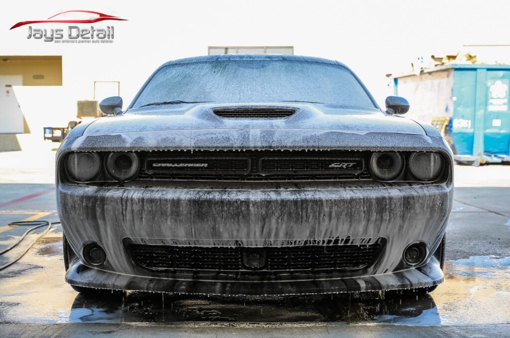 Black Dodge Challenger covered in white foam, being washed outdoors.