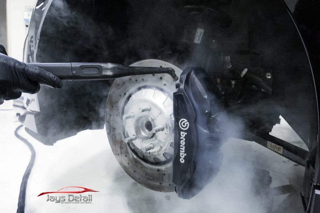 Black-gloved hand cleaning a car brake with a steam cleaner, producing a cloud of vapor.