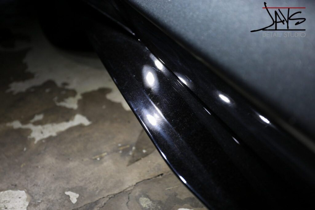 Close-up of a black car's side skirt with scratches, reflecting light on a concrete surface.