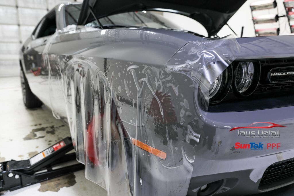 A gray Dodge Challenger with clear protective film being applied to the front bumper in a garage.