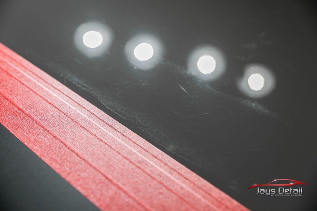 Black car paint reflecting four bright lights, with red tape along the bottom edge, detailing work.