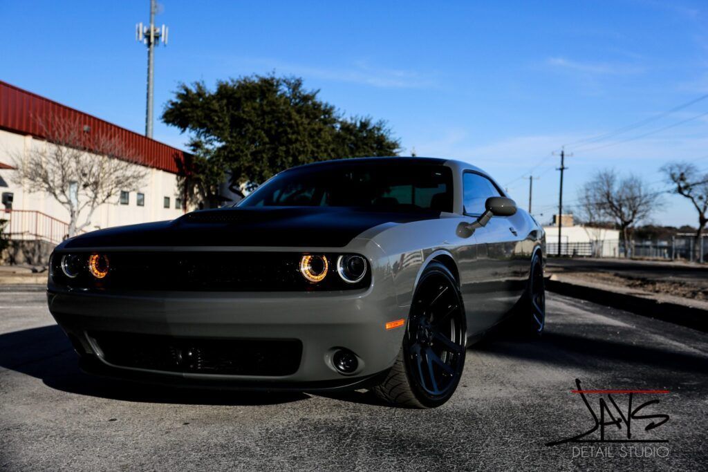 Gray Dodge Challenger with black hood and wheels, parked on asphalt under a blue sky.