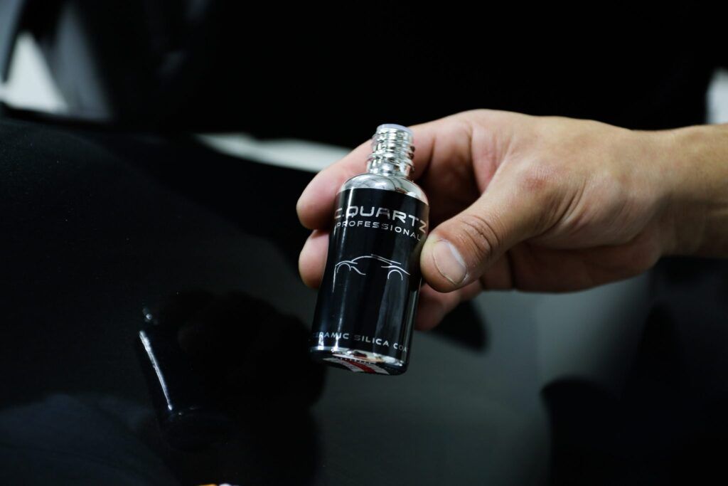 Hand holding a black bottle near a car's glossy surface, presumably applying a detailing product.