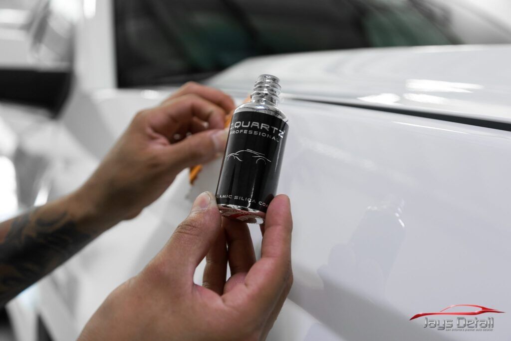 Hands holding a dark bottle on a white car, likely applying a detailing product.