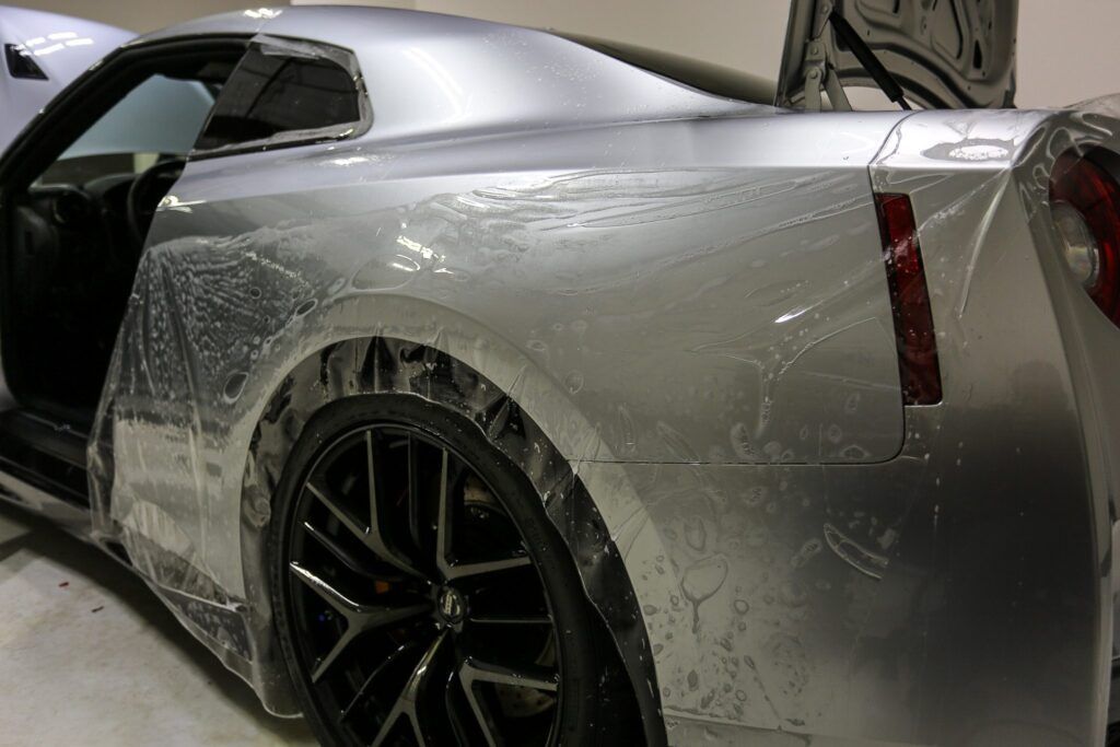 Silver sports car partially covered in clear protective film. Rear tire and open door visible.