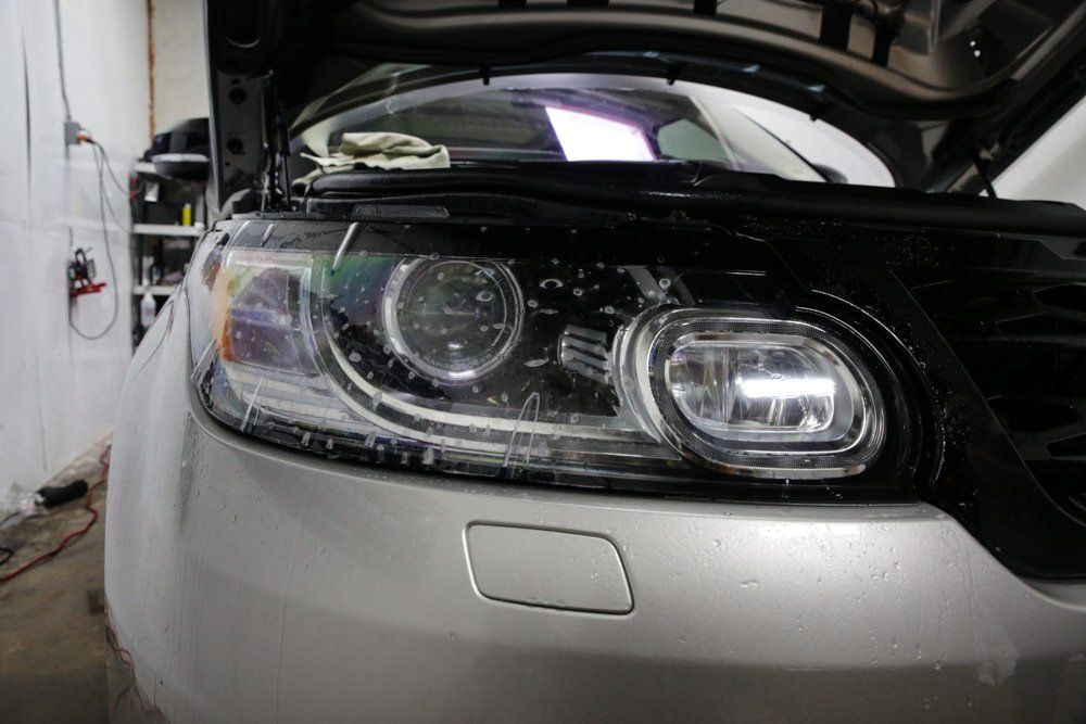 A silver car with the hood open. Front headlight is being cleaned with a spray bottle in an indoor setting.