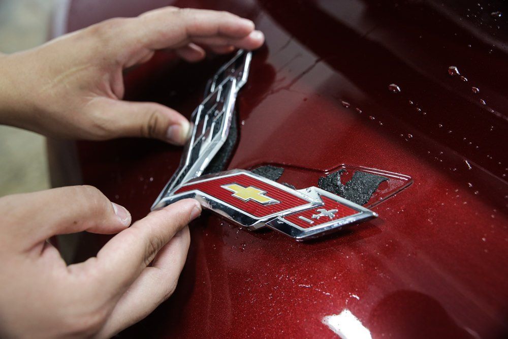 Hands placing a chrome and red car emblem on a shiny maroon surface.
