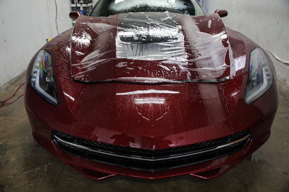 A red Corvette covered with clear protective film in a garage setting.