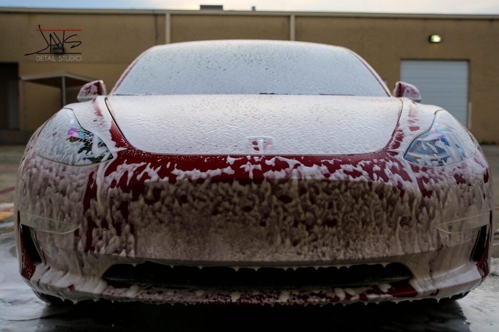 Red Tesla car covered in white foam at a car wash.
