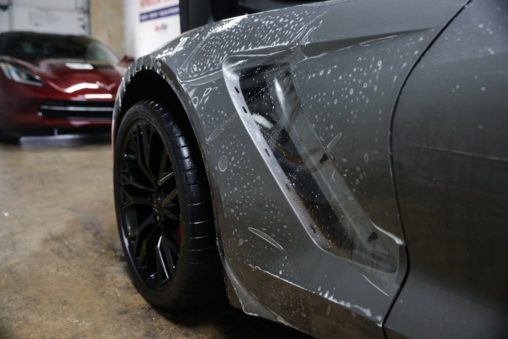 Gray sports car with protective film applied to the front fender, black wheel, and red car in the background.