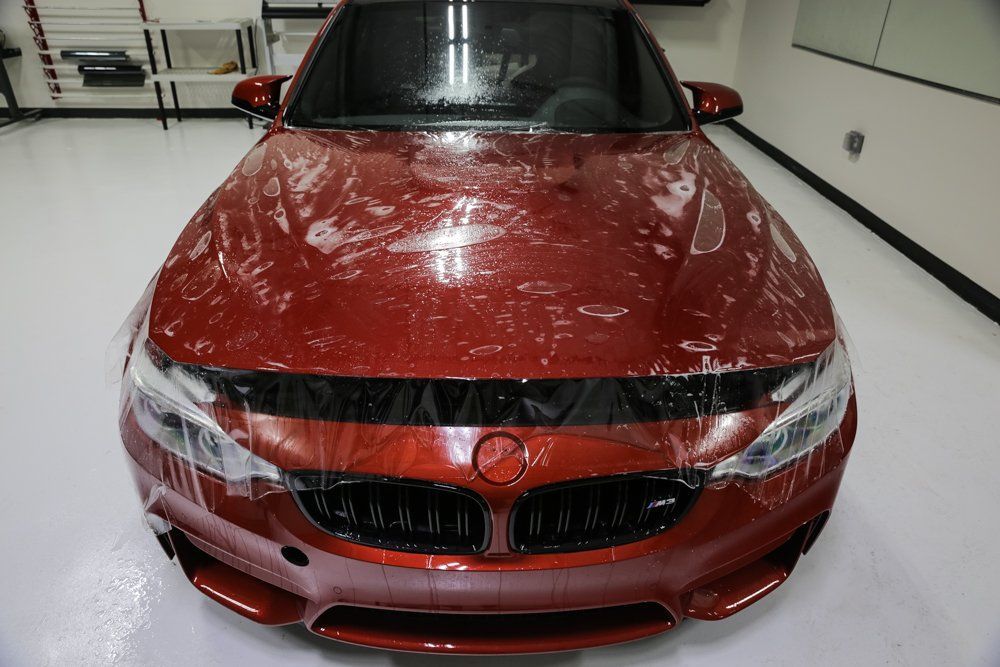 Red car hood covered with protective film, inside a brightly lit garage.