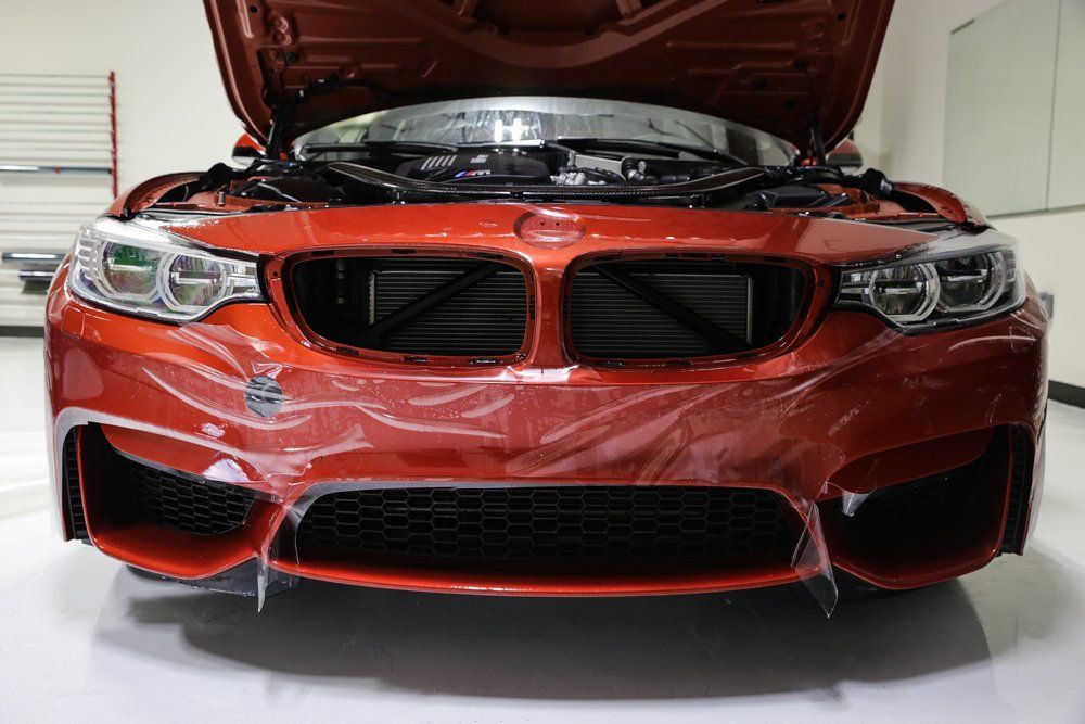 Orange BMW with open hood, front view, protective film on bumper.