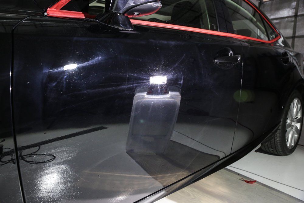 Black car door being polished, showing swirl marks and reflection of a polishing tool.