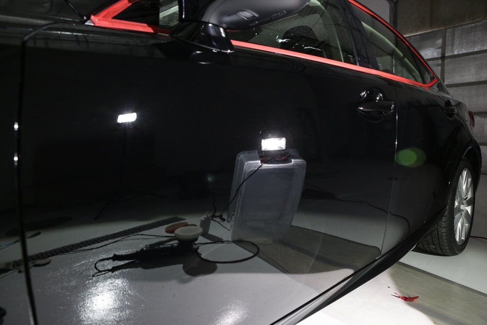 Black car being polished in a garage, showing reflections, with a buffer and work light.