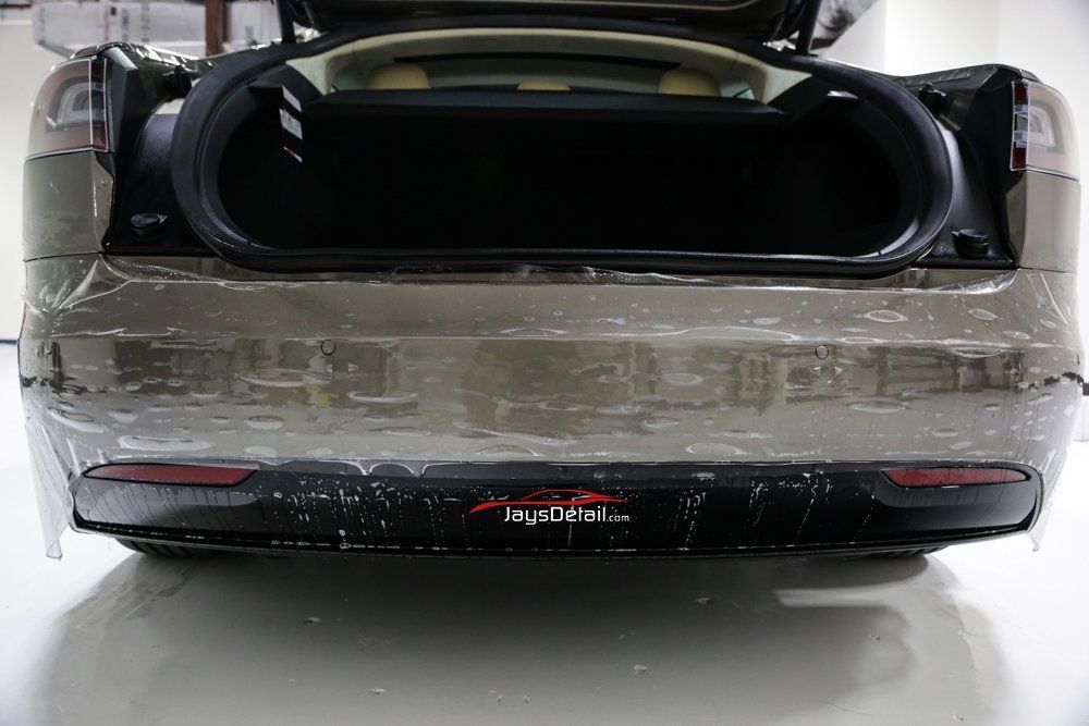 Rear view of a Tesla with the trunk open, showing a black interior and clear protective film on the bumper.