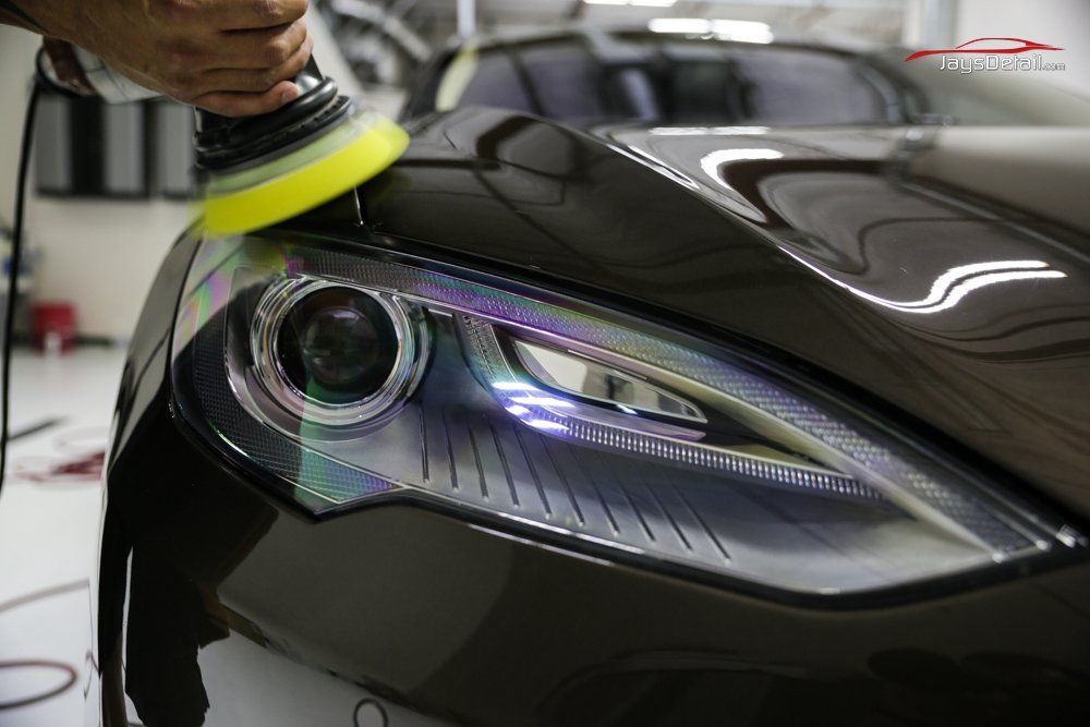 Person polishing car headlight with a yellow buffer in a garage, headlight reflecting light.