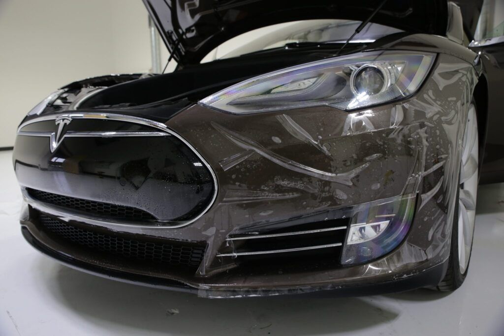 A dark Tesla car with a protective film on the front, hood open, in a white-walled room.