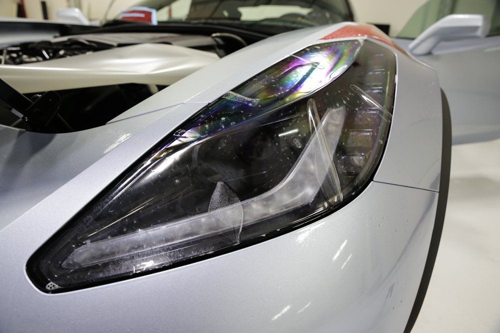 Silver sports car with a clear, angled headlight, hood open, inside a bright room.